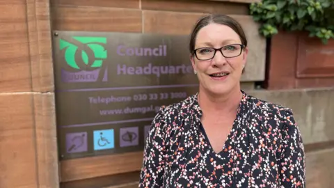 A woman with dark, pulled back hair and glasses in a patterned blouse stands in front of Dumfries and Galloway Council headquarters
