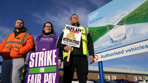 Two men and a woman stand on a picket line with a large Scottish Water billboard - featuring a glass of water against a scenic backdrop - to their right.