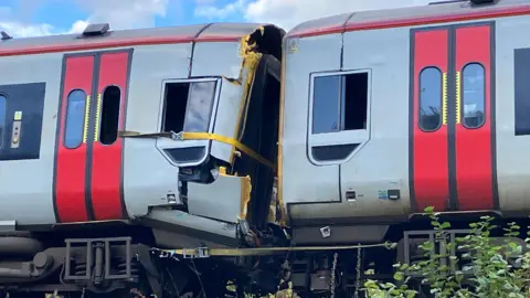 A crumpled section of a train carriage against another slightly bent out of shape carriage. The badly damaged section is being held together by yellow ratchet straps 