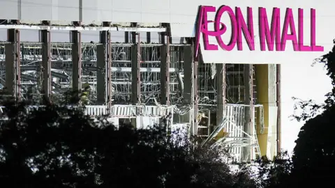 A damaged wall is seen on the side of Aeon Mall Kumamoto. The words Aeon Mall are seen in bright pink letters.