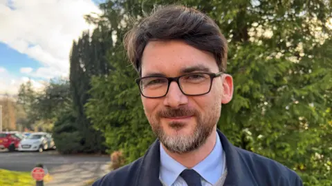 Markus Campbell-Savours is standing in front of a leafy bush and smiling slightly at the camera. He has short black hair and dark, square glasses. He is wearing a blue jacket, blue tie and blue shirt.