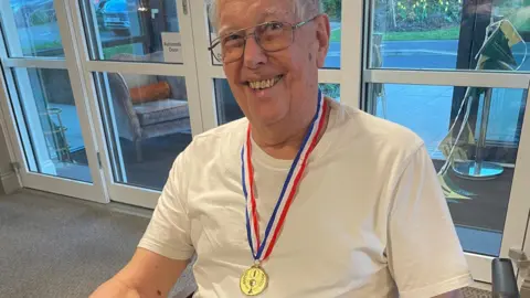 BBPR Leonard Symonds, 88, wearing a white t-shirt with a medal around his neck. He is wearing glasses and is smiling at the camera.