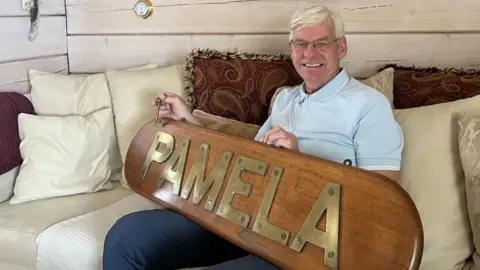 Barour Jon Grimsson A man with white hair smiles as he sits on a cream-coloured sofa while holding a large wooden name plate with gold capital letters spelling out the name "Pamela". He is wearing a light blue polo shirt, dark blue trousers and glasses.