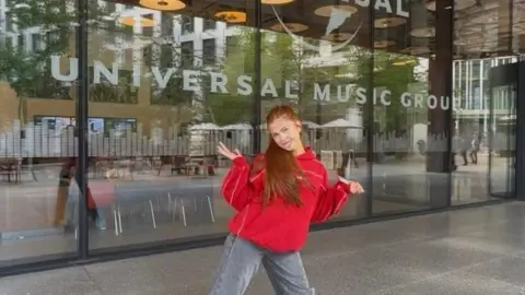 Family handout Sophie is standing in front of the Universal Music Group building. She has long ginger hair in a ponytail and is wearing a red hoodie and grey jeans. 