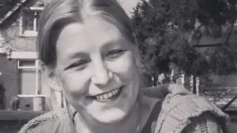 Family Handout A black and white picture of Dawn Sturgess smiling at the camera, wearing a t-shirt, cardigan and a necklace. She is sitting on the grass in front of a house and a tree.