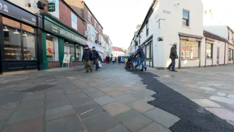 BBC/David Reeves A patio pedestrianised shopping street with shops on either side and people walking.