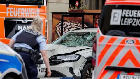A police officer standing in front of a white car with a smashed windscreen surrounded by fire trucks