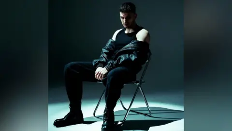 SICKO.RAW A man with dark hair is sitting on a chair wearing a black vest, black trousers and black boots. He has a leather jacket halfway off, and is staring at the camera. There is a spotlight on him which is creating a shadow on one side of his face