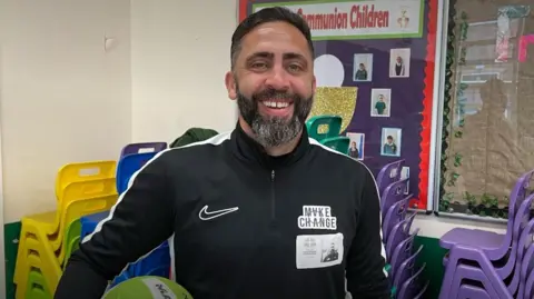 Jay Popat smiling at the camera. He is dressed in a dark sports kit. Two notice boards and some stacked chairs can be seen behind him.