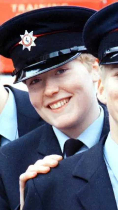 Fleur Lombard looking straight at the camera, smiling. She is wearing a firefighter navy hat with a badge at the front and a navy suit with pale blue shirt and navy tie. 
