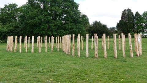 65 wooden poles standing upright in the ground. The ground is grass and the area is surrounded by green trees.
