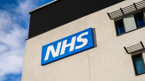 Bright white NHS lettering sits on a blue background, sitting high on the side of a grey building with blue sky visible behind it. 