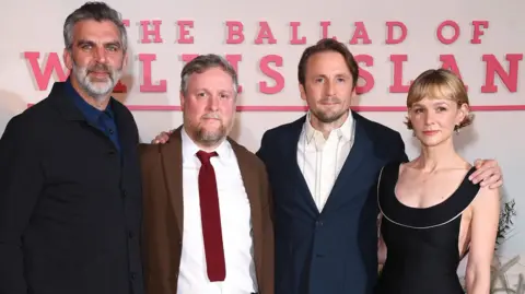 Getty Images Left to right: James Griffiths, Tim Key, Tom Basden and Carey Mulligan against a pink photo call background for the film. Griffiths wears a black jacket and navy blue shirt. He has greying hair and beard and blue eyes. Key wears a white shirt, brown suit jacket and red tie and has short hair and a goatee beard. Basden wears a white shirt with a closed navy suit jacket and has short hair and stubble. Mulligan wears a scooped-neck black dress and has cheekbone-length blonde hair. 