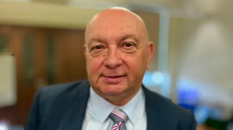 Martin Giles/BBC Carl Smith, leader of Great Yarmouth Borough Council. He is looking directly at the camera and smiling. He is wearing a pale blue shirt with a navy blazer over the top and a grey patterned tie.