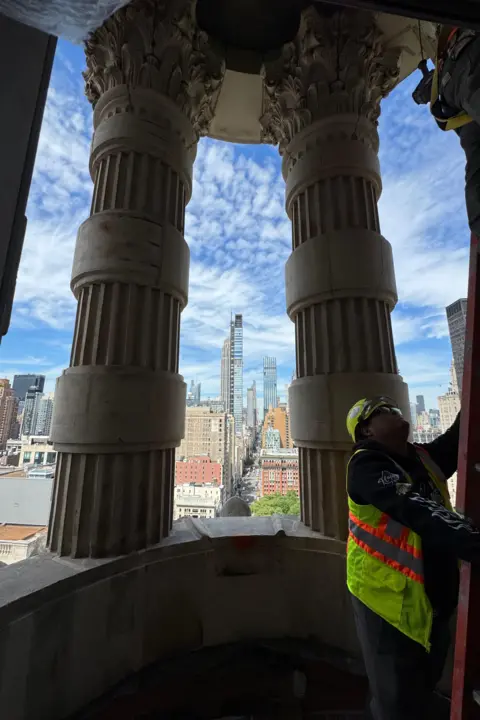 Grace Eliza Goodwin Image of construction worker in yellow vest with a ladder on a balcony with large columns and a view of the Manhattan skyline