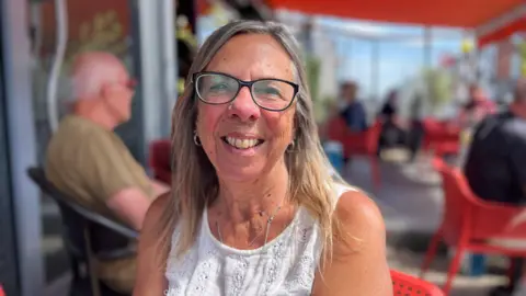 A woman sat outside a cafe wearing a white sleeveless top with black glasses. She is long grey and blonde hair and is smiling at the camera 