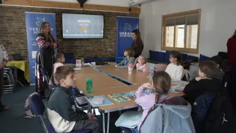 A class of young children sat on a desk gathered around a smart screen. Two female teachers are standing at the front of the class.