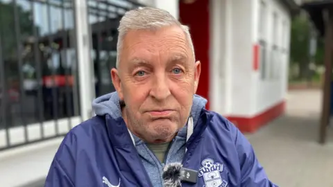 Chris Jewell has short grey white hair and blue eyes. He wears a blue anorak.