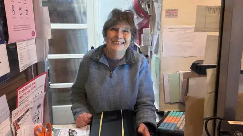 Kate Bradbrook/BBC Jenifer Fell stands in the Post Office counter inside her home. She is smiling at the camera. 