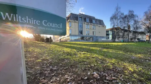 A picture of Wiltshire Council's headquarters in Swindon, as taken from afar. The building is standing in the middle of a large grassy area, and is a sand coloured building with lots of windows. In the foreground there is a Wiltshire Council sign. 