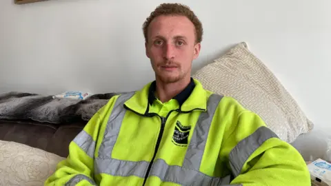 A man sat down on a sofa, wearing a high-viz yellow jacket.
