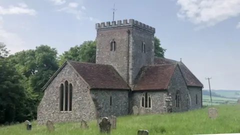 The Valley Parish A small grey stone church on a grassy hill, with large green trees in the background and a grassy graveyard in the foreground