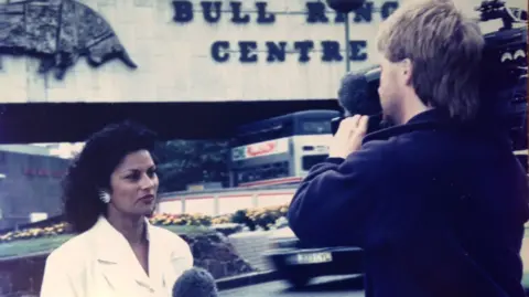 Anita Bhalla A female news reporter in a white outfit is being filmed by a male camera operator outside a building. Behind them is a sign that reads “Bull Ring Centre” with a metal bull sculpture above it, and traffic and flowerbeds in the background.
