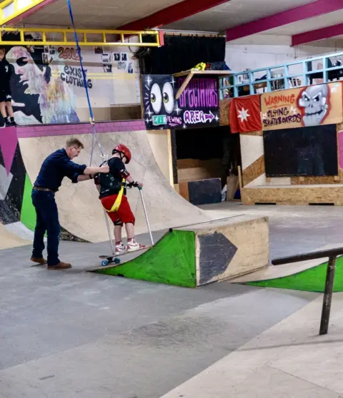 Exist Skatepark A man holds onto a harness attached to a boy on a skateboard. He is wearing a red helmet and a black top with a colourful top on. He is going up a skateboard ramp wearing red shorts and red and white trainers. A skateboard ramp can be seen in the background.