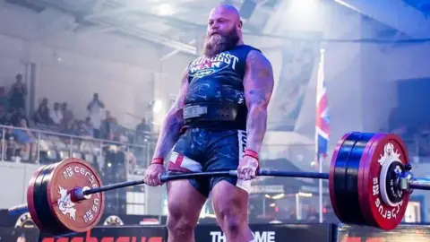 Ultimate Strongman A man in a dark leotard, with a bald head and long brown beard is holding a huge weight just above his knees. His leotard has the words "UK Strongest Man Live" printed on it.