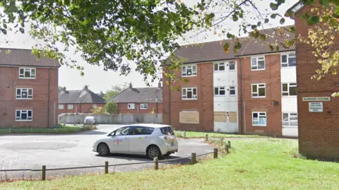 Several windows are visible in a three-storey building on the right and three sets of windows can be seen in a three-storey building on the far left. A car is parked in the foreground.