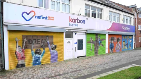 Karbon Homes Children are painted on the shutters of Karbon Homes' Skills Hub and neighbouring NEFirst Credit Union. They show the kids with their hands up in the air in front of a sign reading DREAM, climbing a tree, standing in front of houses and playing below a yellow writing reading WE MAKE STANLEY.
