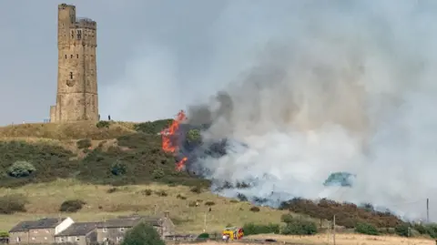 A fire creeps up a hill towards a tower. Smoke can be seen billowing into the sky with a fire engine parked at the foot of the hill and one or two firefighters visible near the start of the fire.
