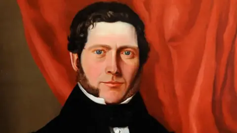 Guernsey Museums & Galleries A painting of William Le Lacheur, he is smiling with his mouth closed and there is a red curtain behind him. He has long brown mutton chops and dark hair.