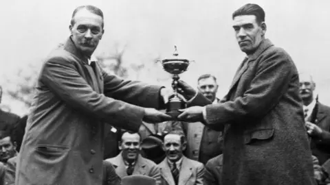 Getty Images A black-and-white photograph shows a trophy presentation ceremony, connected to the early years of the Ryder Cup. In the foreground, Samuel Ryder and George Duncan stand in long overcoats facing the camera while jointly holding a large trophy between them.