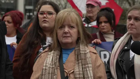 Donna Whyte stands outside the court, wearing a light brown coat, checked brown scarf and blue t-shirt. Her face is sombre and she is flanked by family members and protestors.