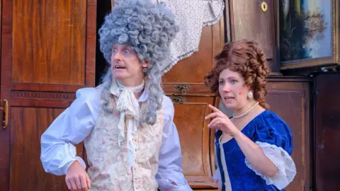 Chris Kelly Ben Kernow and Eleanor Toms are wearing large wigs and full costumes. Ben has a white shirt and cream patterned waistcoat and Eleanor is in a dark blue top with lace edging.