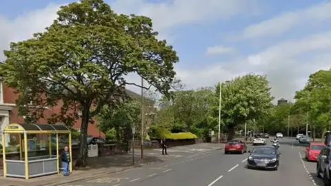 Google A wide tree-lined road where a man is stood at a bus stop. A number of cars and a motorbike are travelling along the road.