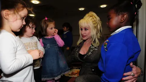 Getty Images Dolly Parton crouches down and speaks to several children. She has her arms around two of them.