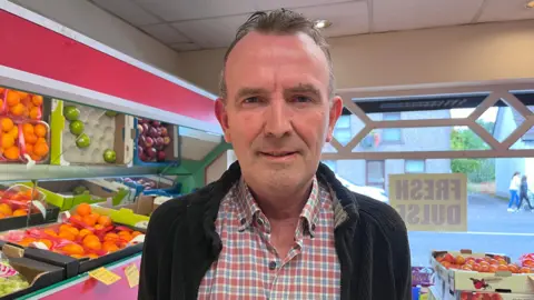 John Devlin in his fruit and veg shop smiling at camera. He is wearing a red and blue chequed shirt with as a black jacket on. 