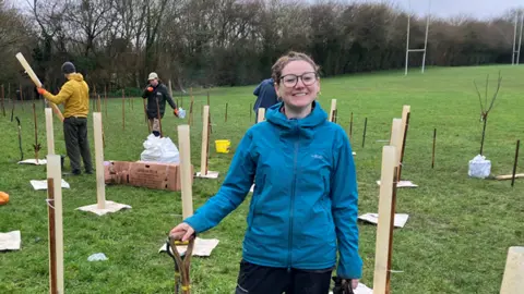 A woman holding a spade stand on a patch of grass. Around her are a team of people planting trees. 