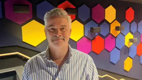 Photograph of Colin Lawrence from the hospital cinema charity MediCinema. He is pictured in front of brightly coloured hexagons inside the hospital.