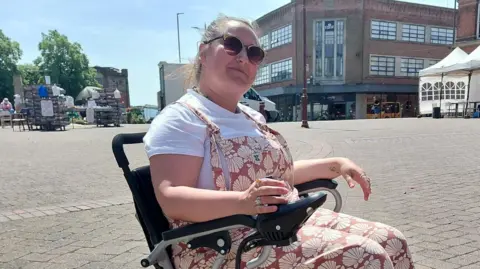 A brown haired woman wearing sunglasses and a white t-shirt with pink dungarees in a black motorised wheelchair.