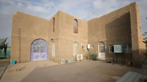 A house that appears to be made of concrete, with a flat roof and arched windows. There are two intricately patterned metal doors - one is lilac and the other is white. There is a courtyard in the foreground.
