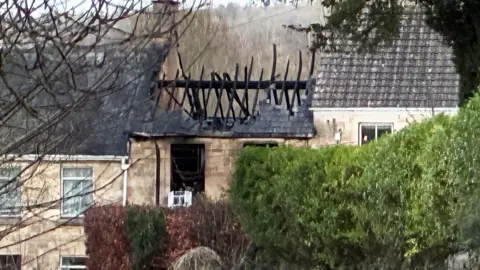 The charred shell of the destroyed home following a fire. The roof has collapsed and only the burnt wooden beams are visible. The glass windows have been smashed and are completely blackened from smoke. The surrounding homes on the terrace are untouched.