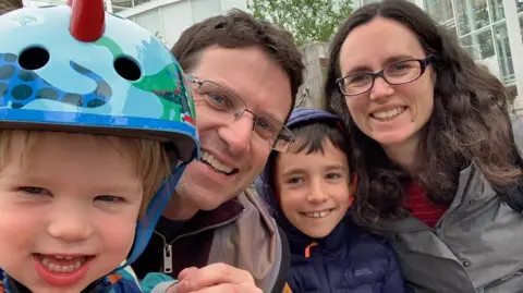 Dr Alexis Gilbert A family of four are standing outside a large building and smiling at the camera.  One young child is wearing a cycling helmet.  Both parents are wearing glasses.