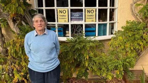 Justine Bayley, wearing a light blue jumper, stands in front of her house with 'No Third Runway' posters displayed in the window