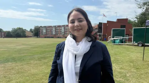 Tom Williams/BBC Hughes Hall By-Fellow, Zahra Joya, is an Afghan journalist, founder of Rukshana Media and activist for the rights of women and girls in Afghanistan. She has long dark brown hair and is wearing a blue jacket, white scarf and top. She is standing on a grass sports field.