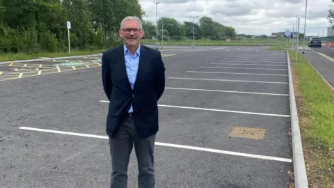 A man standing in an empty car park. He is wearing a suit jacket and shirt and has his arms behind his back.