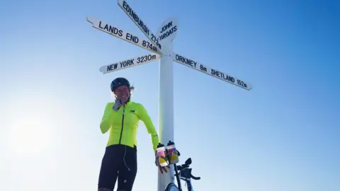 A woman standing in front of the Land's End sign with a bicycle
