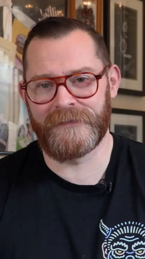 Alex Young looks at the camera. He has short ginger hair with a ginger and white beard. He wears aviator style thick tortoisehell framed glasses and a black t shirt, which has a logo of a viking on it.